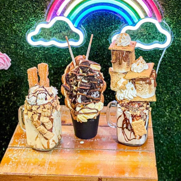 Three decadent La Crepe Og Gourmet desserts with whipped cream, cookies, and churros under a rainbow neon sign.