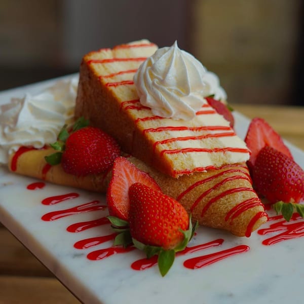A slice of strawberry shortcake with whipped cream, surrounded by crepes, fresh strawberries, and strawberry syrup.