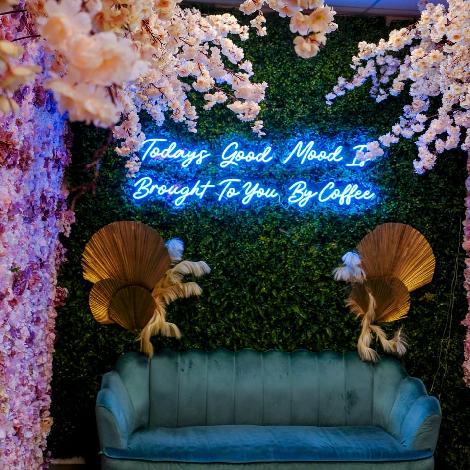 A teal velvet couch against a green wall with a neon sign, surrounded by pink and white flowers.