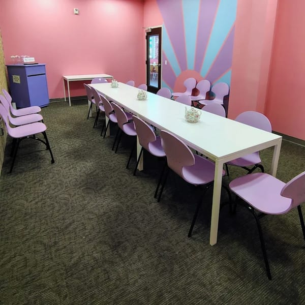 Long white table with purple chairs in a pink room with a sunburst mural.