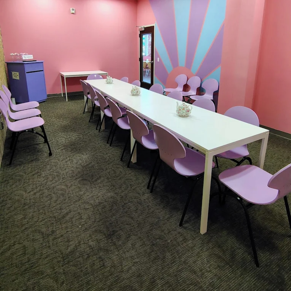 Long white table with purple chairs in a pink room with a sunburst mural.