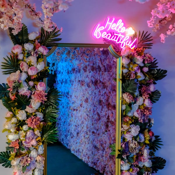 A gold-framed mirror surrounded by pink and white flowers, with a neon "Hello Beautiful" sign above.