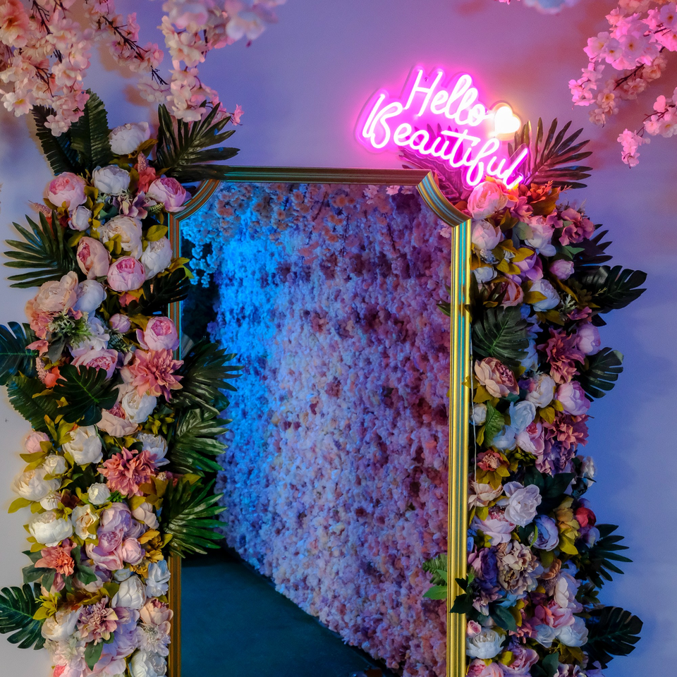 A gold-framed mirror surrounded by pink and white flowers, with a neon "Hello Beautiful" sign above.