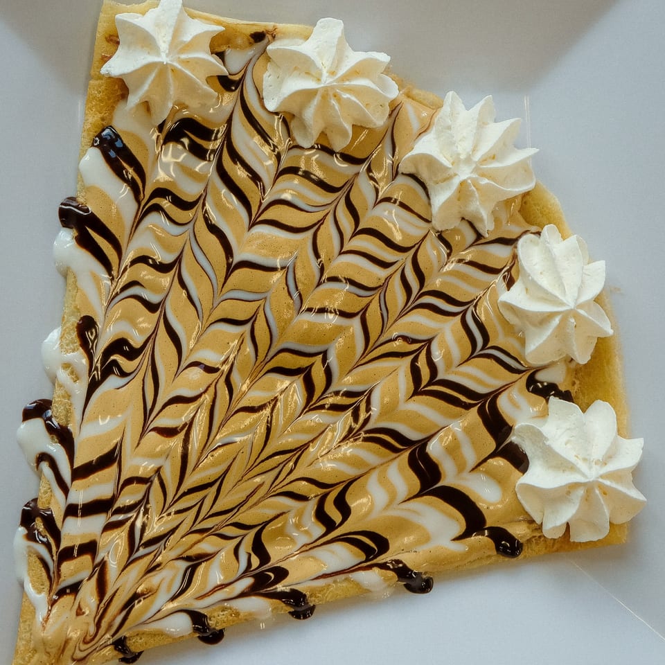 A La Crepe Og Gourmet crepe with chocolate, caramel, and white icing swirls, topped with whipped cream.