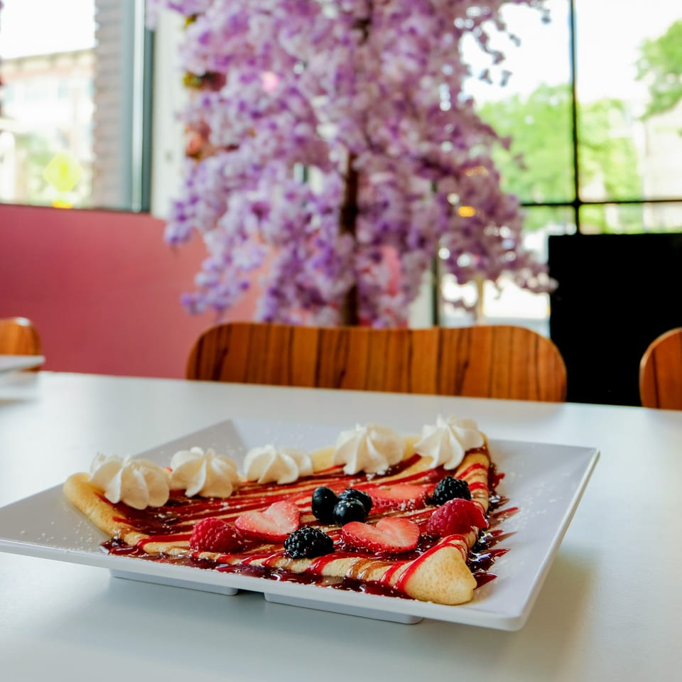 A berry crepe with whipped cream and syrup on a white plate, with a purple floral decoration in the background.